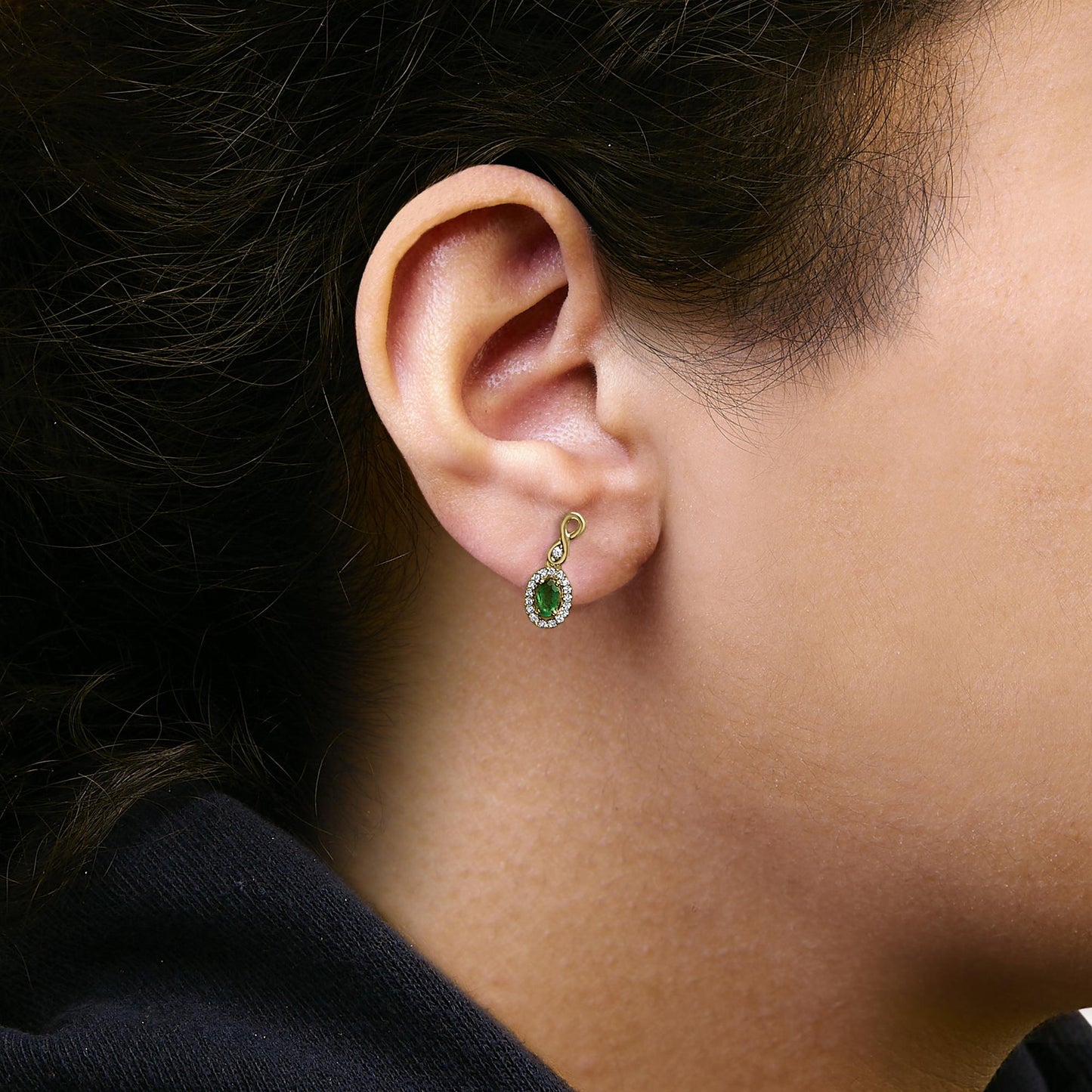 Close-up side view of a yellow metal earring worn on a model, featuring an oval green gemstone surrounded by small white gemstones in a pave setting.