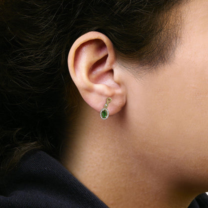 Close-up side view of a yellow metal earring worn on a model, featuring an oval green gemstone surrounded by small white gemstones in a pave setting.