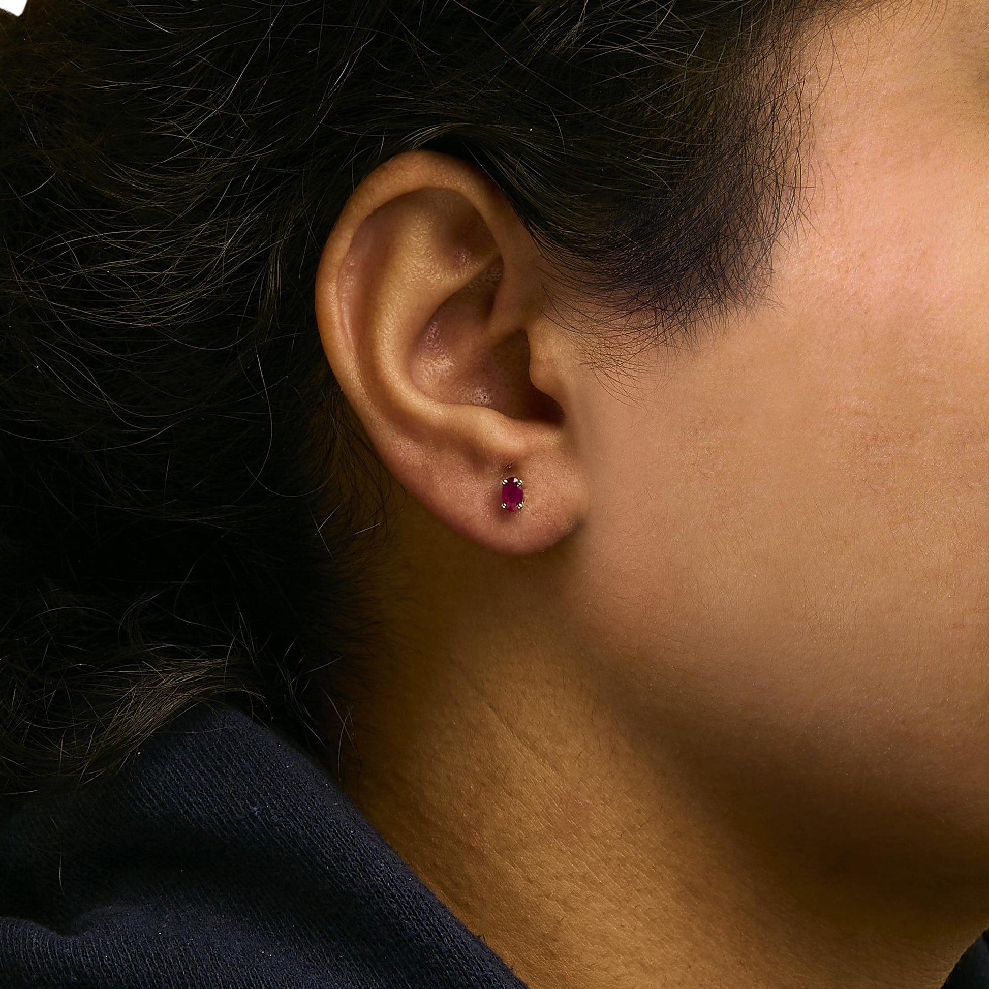Close-up side view of a yellow metal stud earring with an oval red gemstone in prong setting worn on a model's ear.