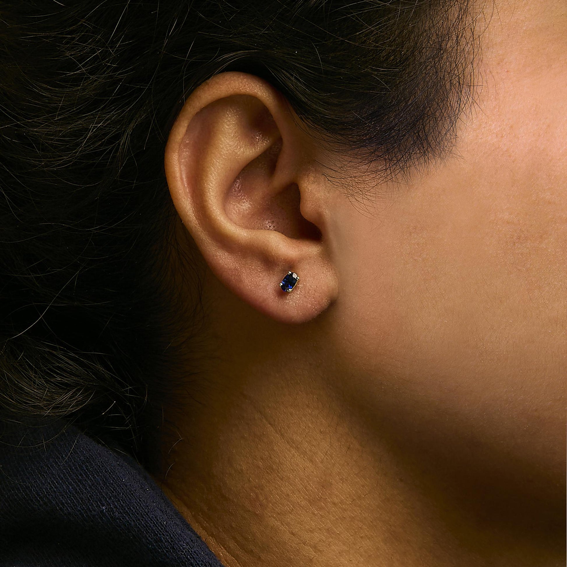 Close-up side view of a yellow metal earring with a small oval blue gemstone in prong setting worn on a model's ear.