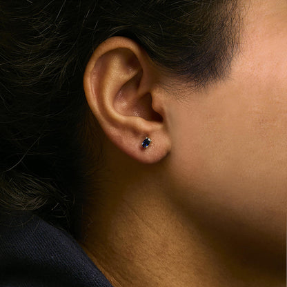 Close-up side view of a yellow metal earring with a small oval blue gemstone in prong setting worn on a model's ear.