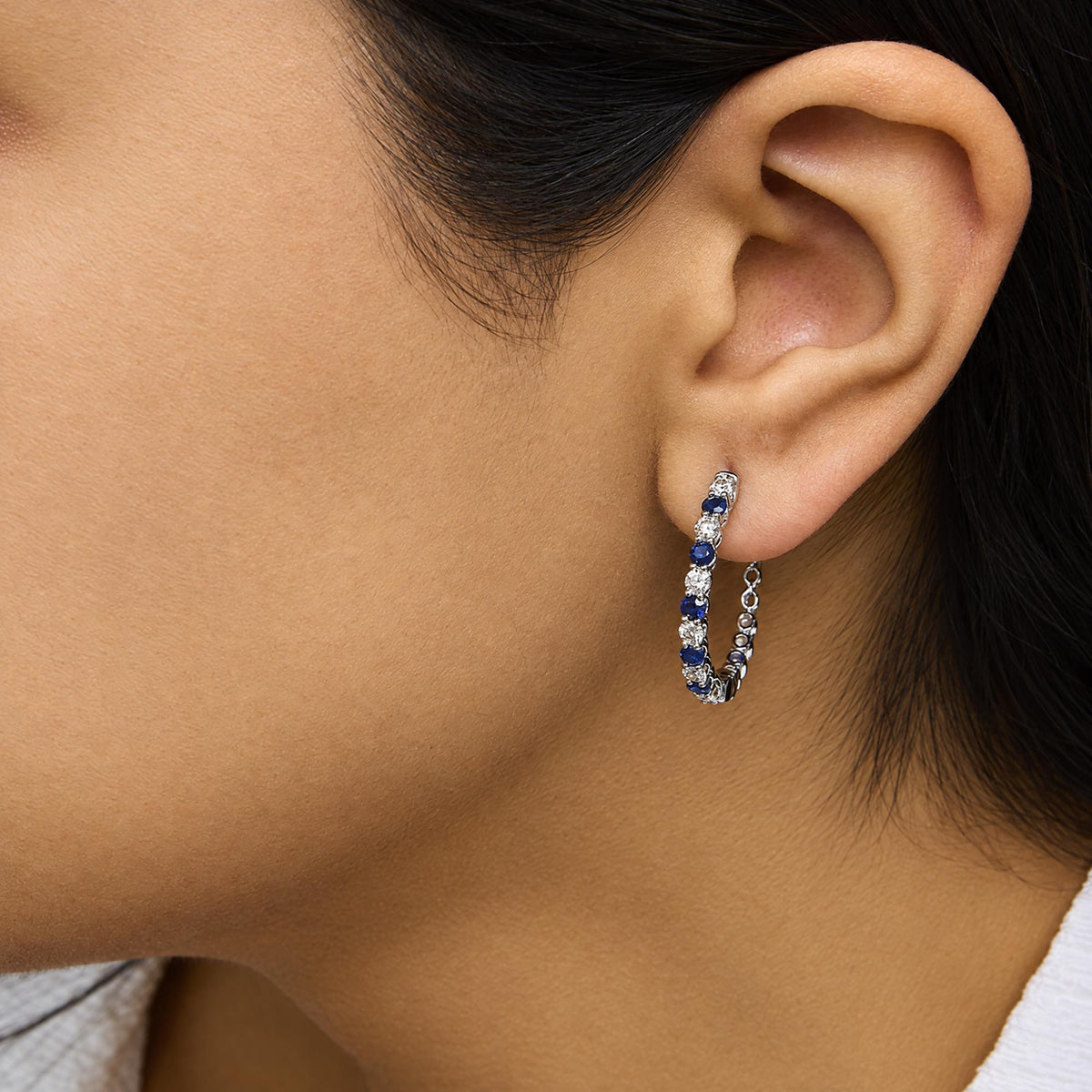 Close-up side view of a white metal hoop earring with alternating white and blue round gemstones in prong settings, worn on a model's ear.