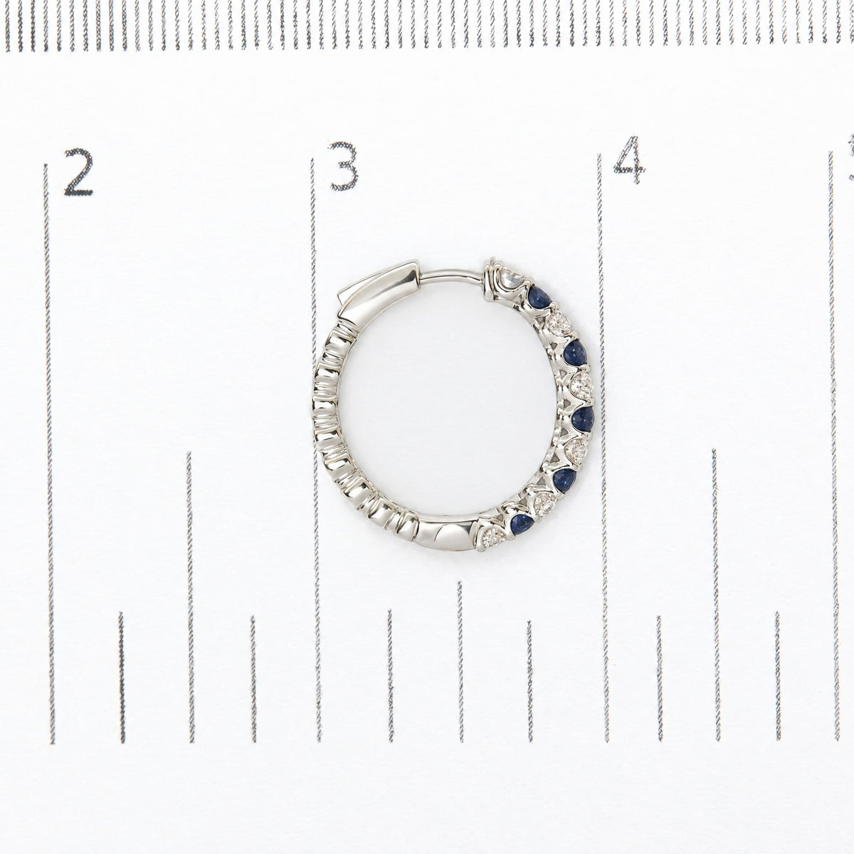 Close-up top-down view of a white metal hoop earring set with round white and blue gemstones in prong settings, shown on a measurement scale.