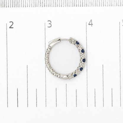 Close-up top-down view of a white metal hoop earring set with round white and blue gemstones in prong settings, shown on a measurement scale.