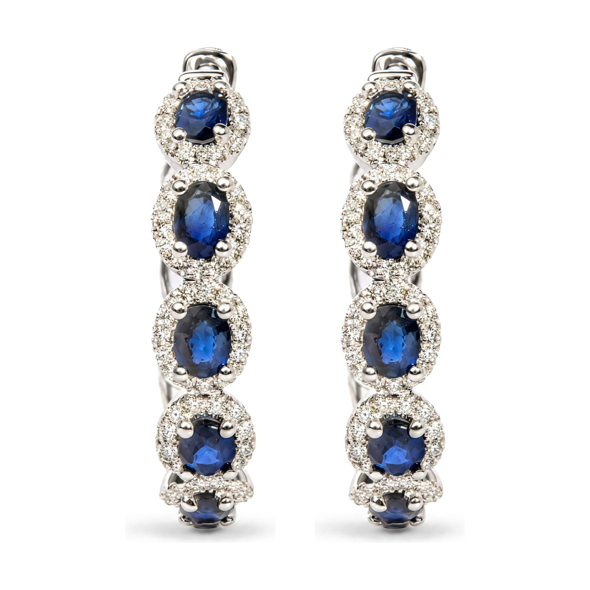 Close-up front view of white metal earrings featuring oval blue gemstones surrounded by small round white gemstones in prong settings.