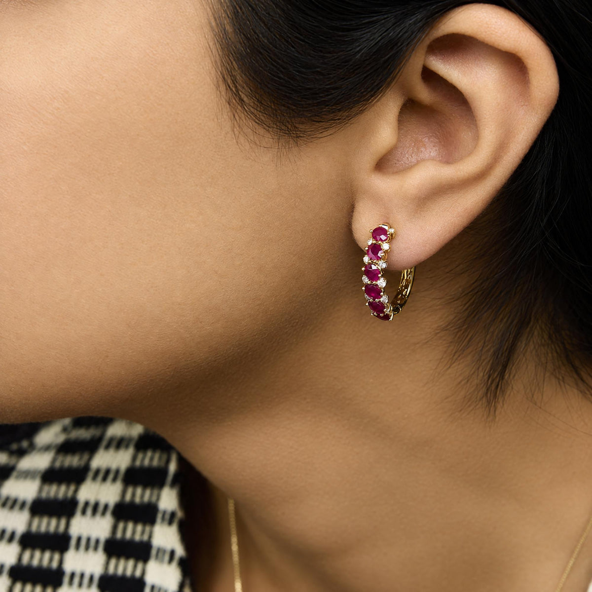 Close-up side view of a model wearing yellow metal hoop earrings set with oval red gemstones and round white gemstones in prong settings.
