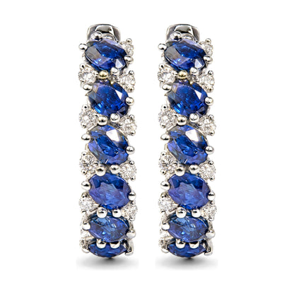 Close-up front view of a pair of white metal earrings featuring oval-shaped blue gemstones and multiple round white gemstones in prong settings.
