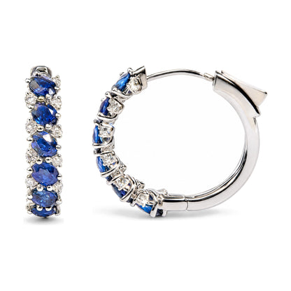 Close-up front and side view of white metal hoop earrings featuring multiple oval blue gemstones and round white gemstones in prong settings, shown on a white background.