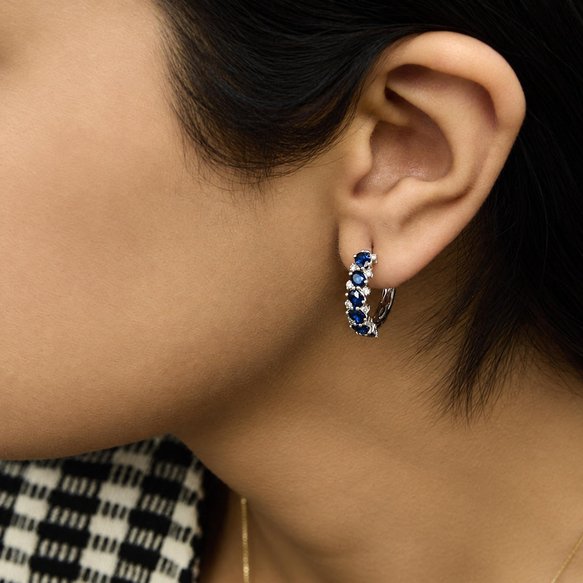 Close-up side angle of a model wearing white metal hoop earrings adorned with oval blue gemstones and round white gemstones in prong settings.