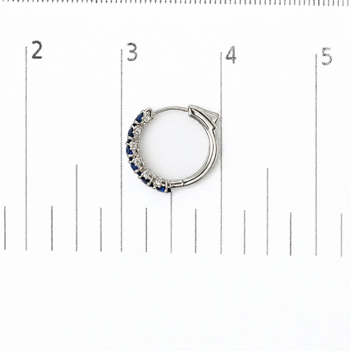 Close-up top view of a white hoop earring displayed on a scale, featuring alternating prong-set oval blue gemstones and round white gemstones along one side.