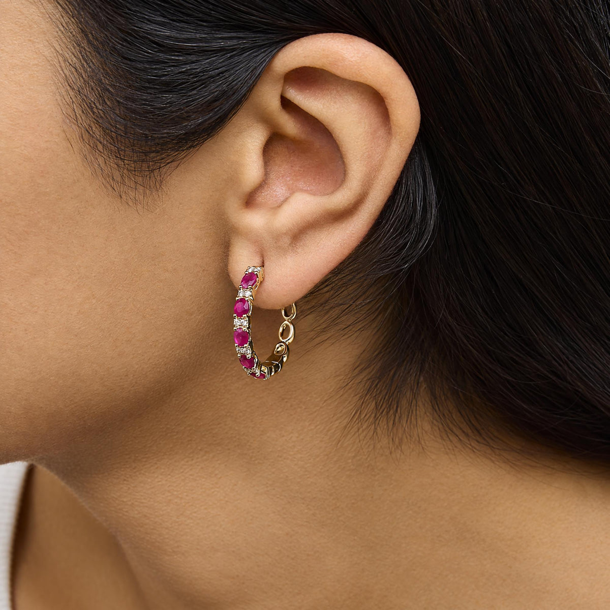 Close-up side view of a model wearing a yellow metal hoop earring with alternating oval red gemstones and round white gemstones in prong settings.