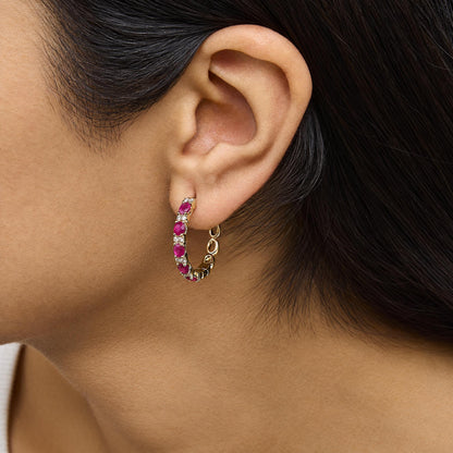 Close-up side view of a model wearing a yellow metal hoop earring with alternating oval red gemstones and round white gemstones in prong settings.