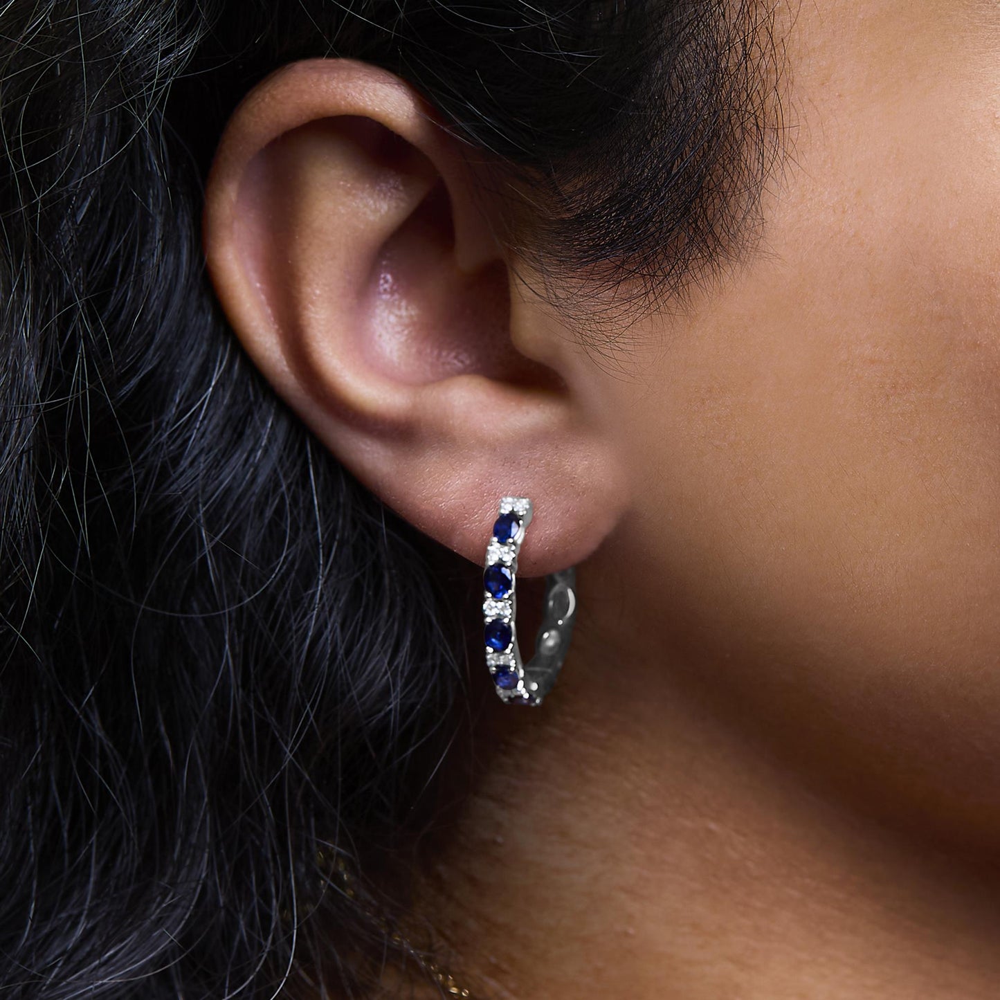 Close-up side view of a white metal hoop earring on a model's ear, featuring alternating oval-shaped blue gemstones and round white gemstones in prong settings.