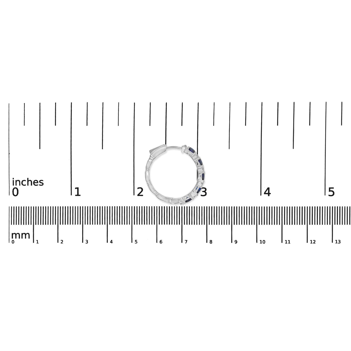 Close-up front view of a white metal hoop earring with alternating small white round gemstones and blue oval gemstones set in prongs, placed on a ruler for size reference.