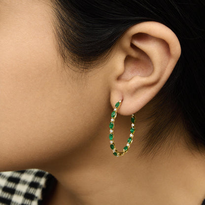 Close-up side view of a yellow metal hoop earring with alternating green marquise-shaped gemstones and white round gemstones, shown worn on a model.