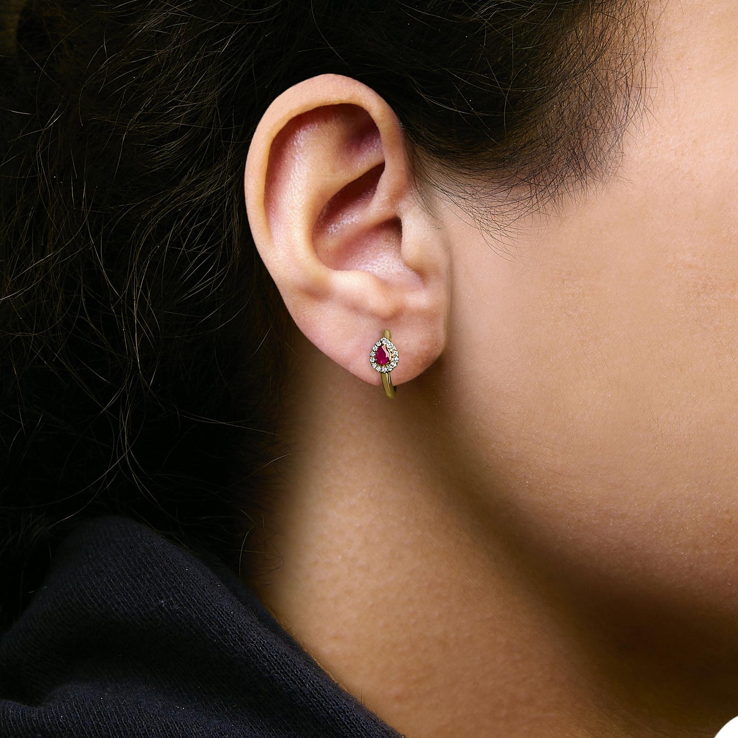 Close-up side view of an earring shown worn on a model, featuring a yellow metal hoop with a pear-shaped red gemstone surrounded by a halo of small round white gemstones.
