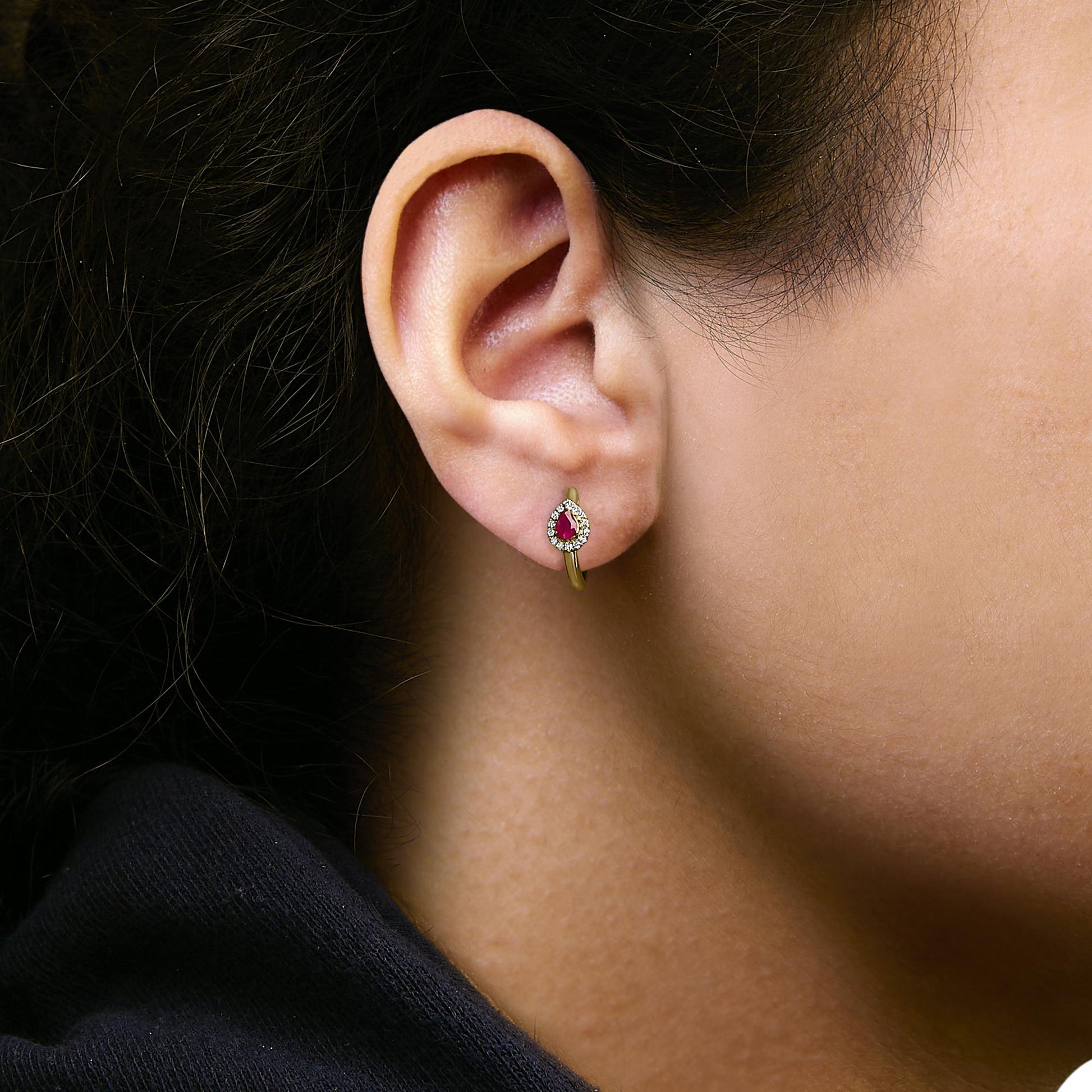 Close-up side view of an earring shown worn on a model, featuring a yellow metal hoop with a pear-shaped red gemstone surrounded by a halo of small round white gemstones.