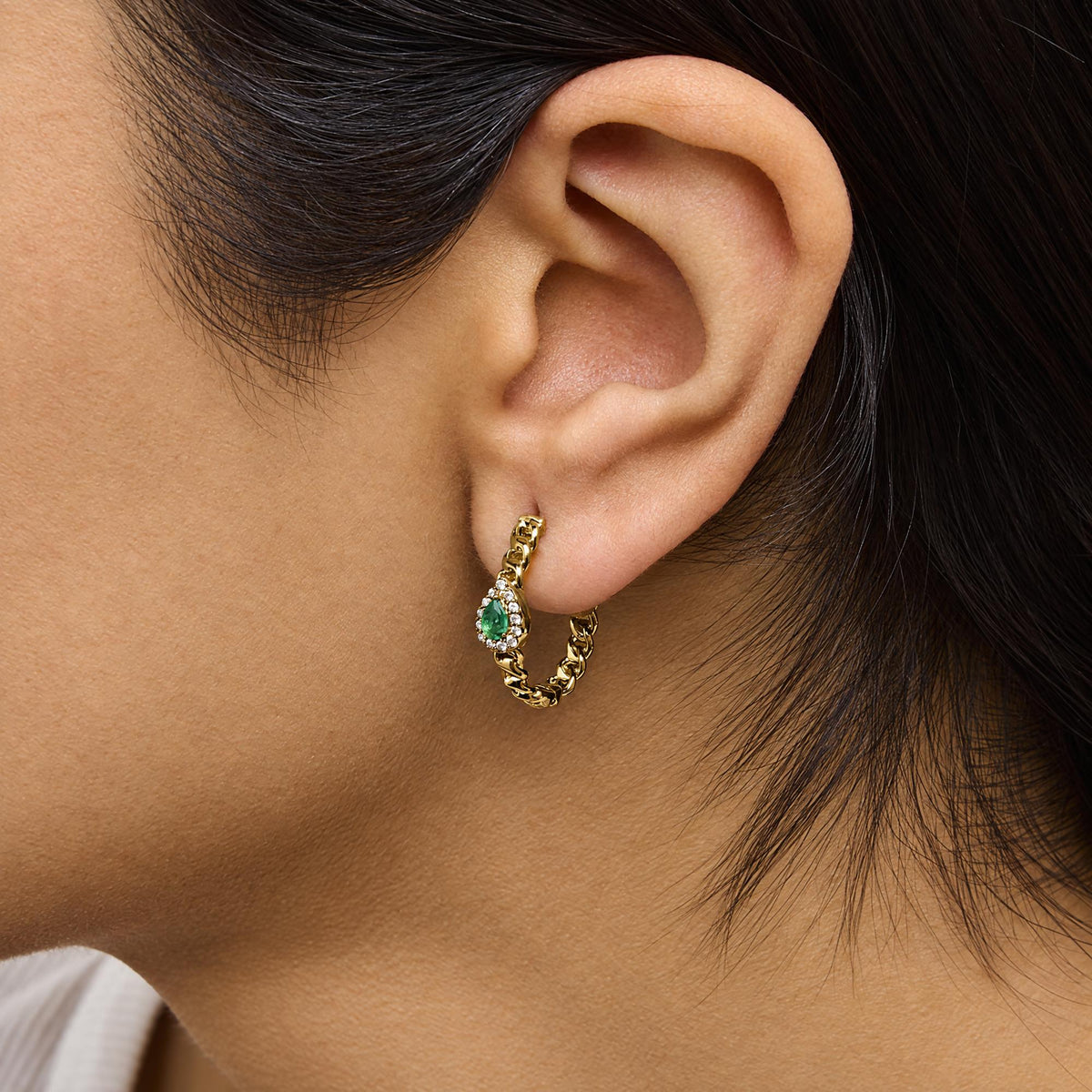 Close-up side view of a model wearing yellow metal earrings with a pear-shaped green gemstone surrounded by round white gemstones in a prong setting, connected by a textured chain link design.