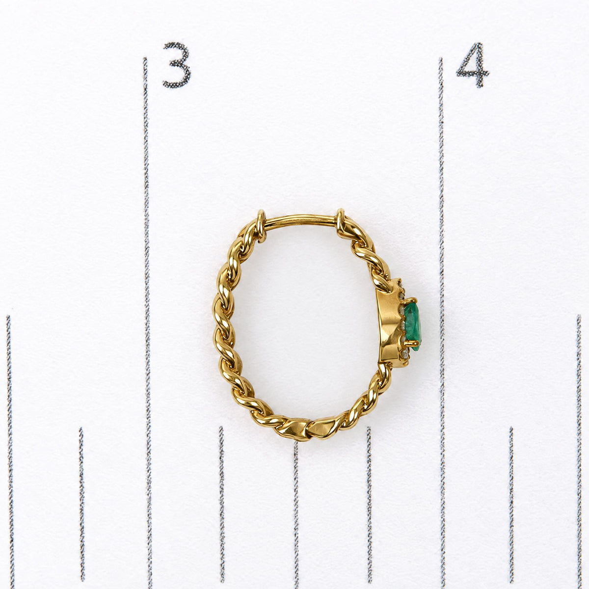 Close-up front view of a yellow metal loop earring with a braided design, featuring a green pear-shaped gemstone and small white round gemstones surrounding it, shown on a scale.