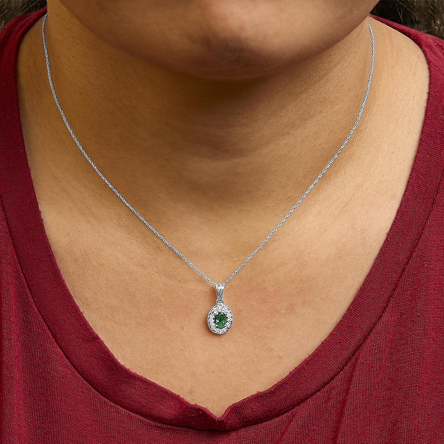 White metal necklace with an oval-shaped green gemstone pendant surrounded by round white gemstones in prong settings, shown worn on a model from a front angle.