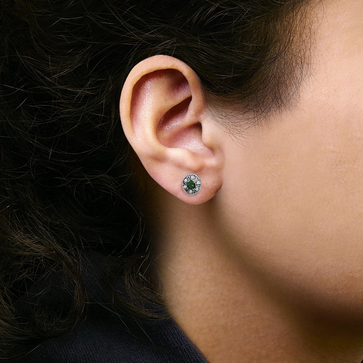 Close-up side angle of a model wearing white metal stud earrings featuring an oval green gemstone center surrounded by a halo of small white gemstones in a pave setting.