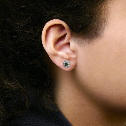 Close-up side angle of a model wearing white metal stud earrings featuring an oval green gemstone center surrounded by a halo of small white gemstones in a pave setting.