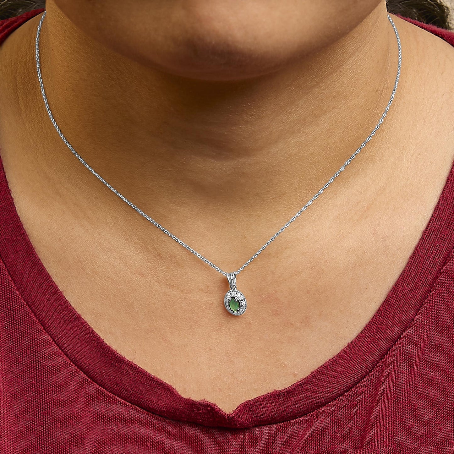 White metal necklace with a small oval pendant featuring a green oval gemstone surrounded by white round gemstones in a pave setting, shown worn on a model from a front angle.