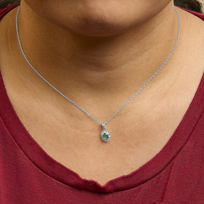White metal necklace with a small oval pendant featuring a green oval gemstone surrounded by white round gemstones in a pave setting, shown worn on a model from a front angle.