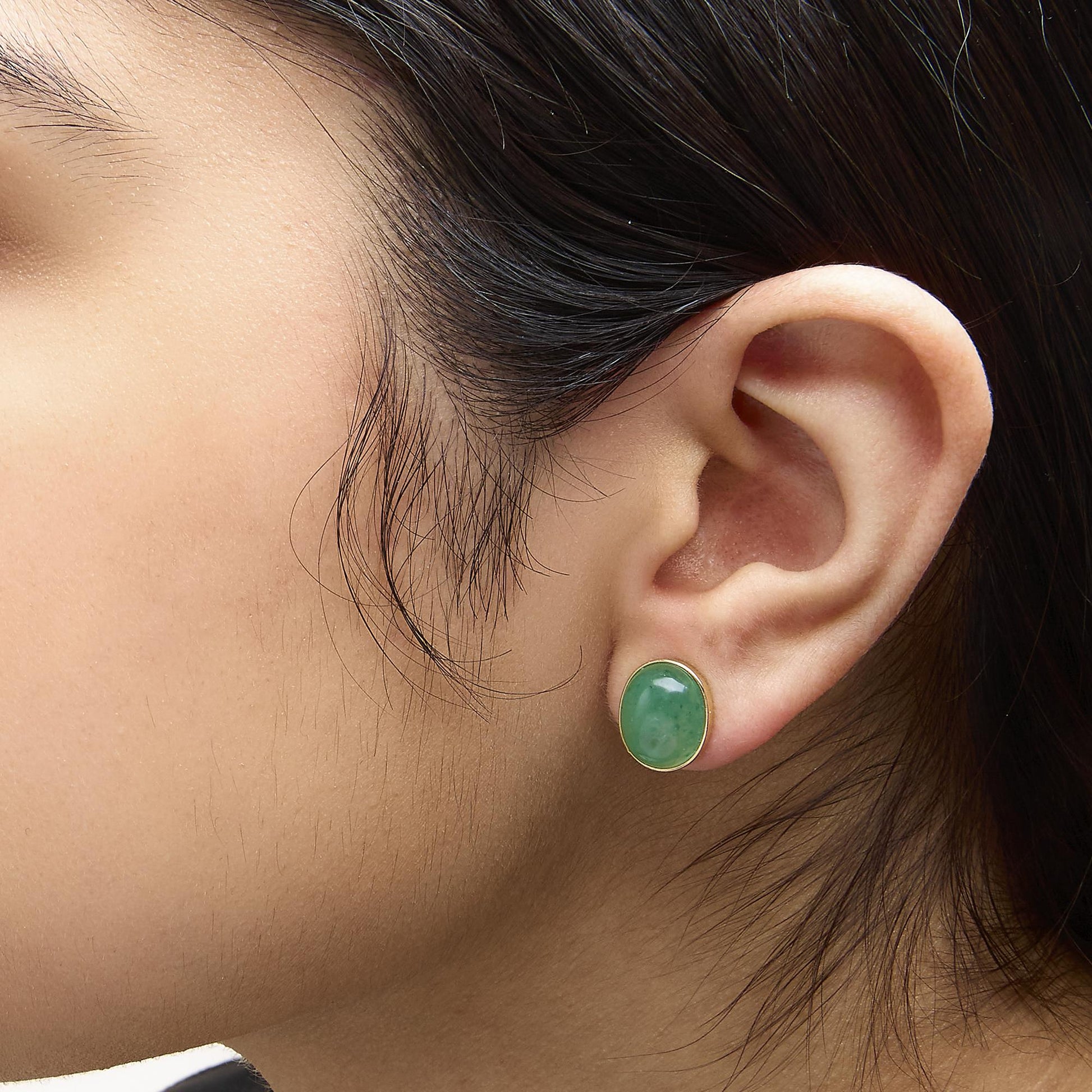 Close-up side view of a model wearing yellow metal stud earrings with oval green gemstones in bezel settings.