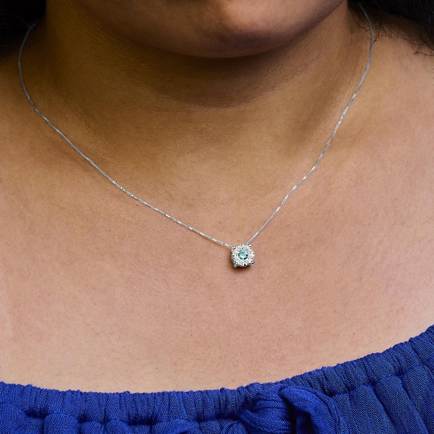 Close-up front view of a white metal necklace on a model, featuring a round green gemstone surrounded by multiple small round white gemstones in a pave setting.