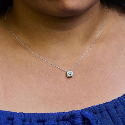 Close-up front view of a white metal necklace on a model, featuring a round green gemstone surrounded by multiple small round white gemstones in a pave setting.