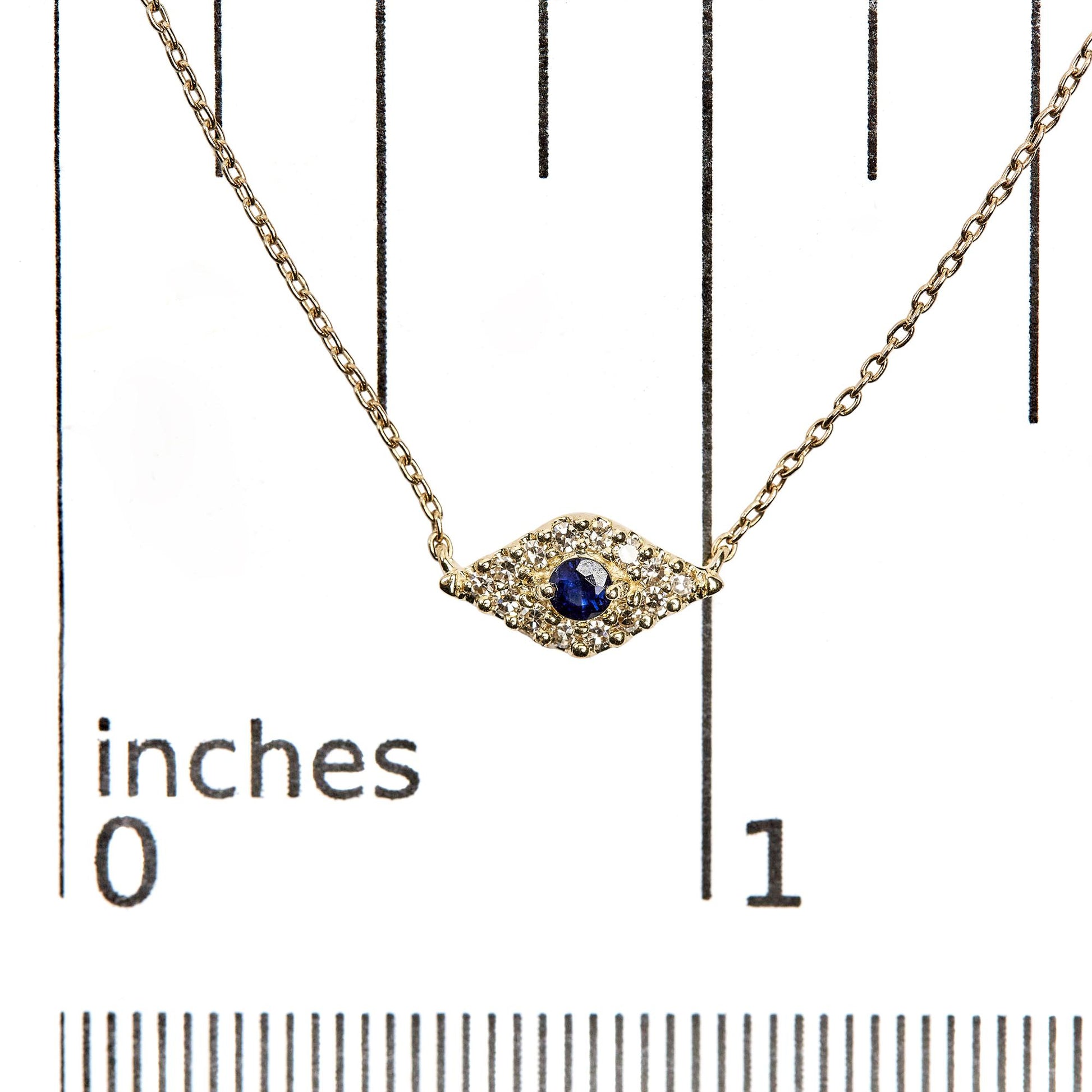 Close-up front view of a yellow metal necklace with a central blue round gemstone in a 2-prong setting, surrounded by multiple small white round gemstones in pave settings, displayed on a ruler for size reference.