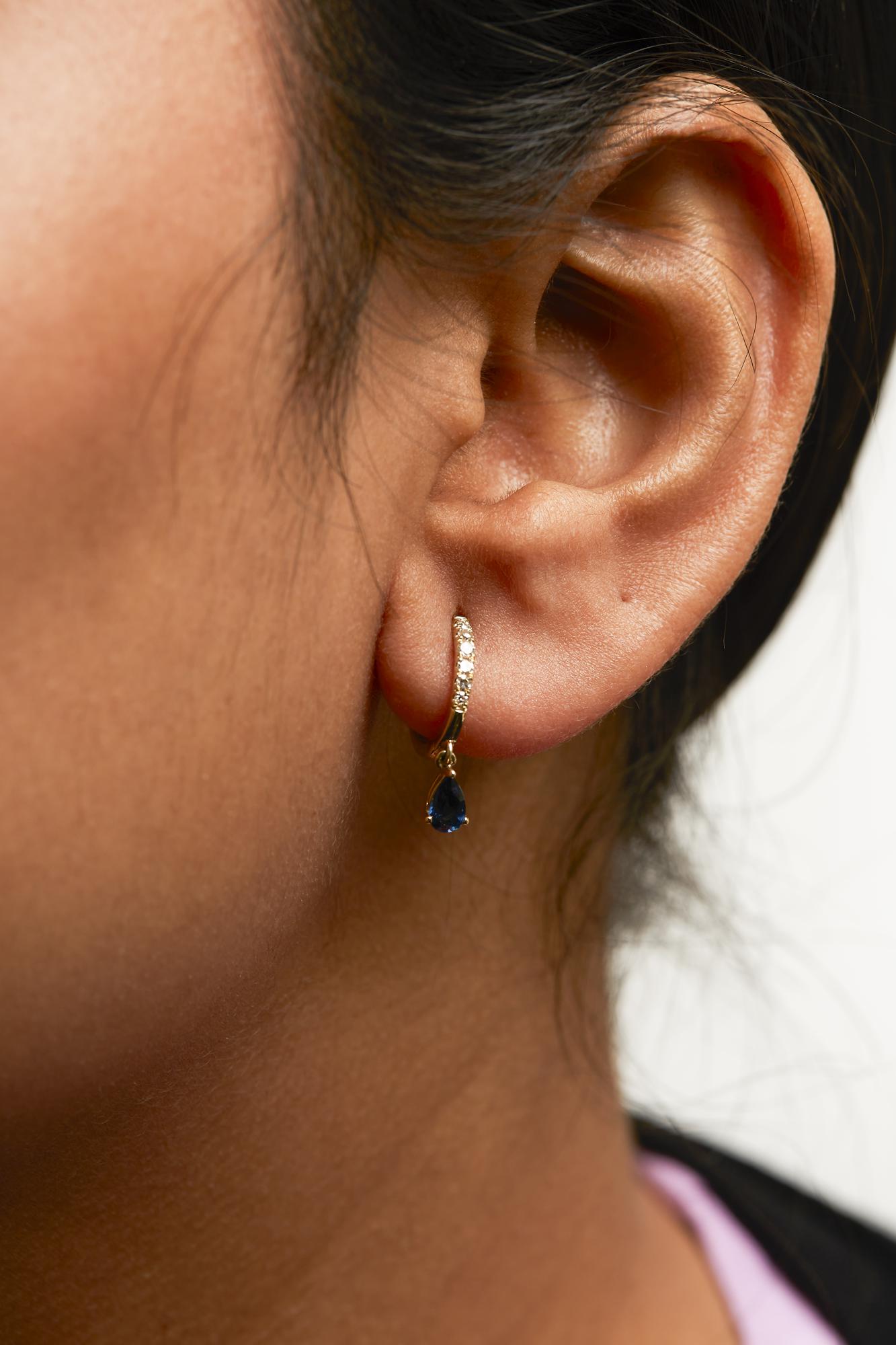 Close-up side view of a yellow metal earring on a model's ear, featuring a small hoop with white gemstones in pave settings and a dangling pear-shaped blue gemstone in a prong setting.