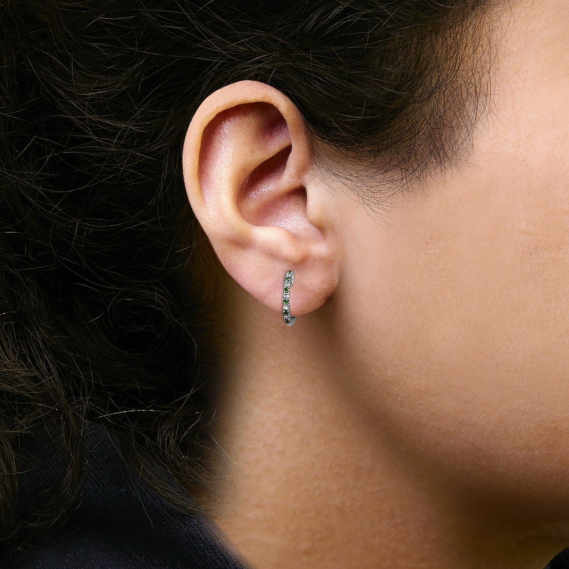 Close-up side view of a model wearing a white metal hoop earring set with round green and white gemstones in prong settings.