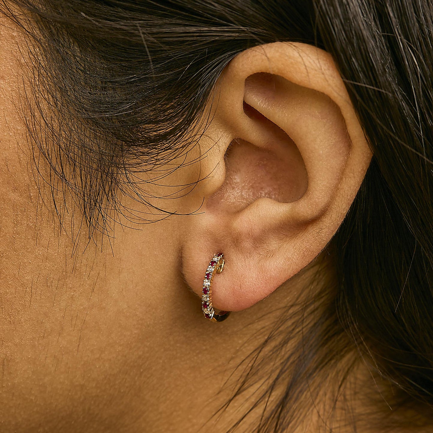 Close-up side view of a model wearing a yellow metal hoop earring set with alternating round red and white gemstones in prong settings.