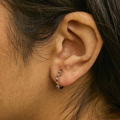 Close-up side view of a model wearing a yellow metal hoop earring set with alternating round red and white gemstones in prong settings.