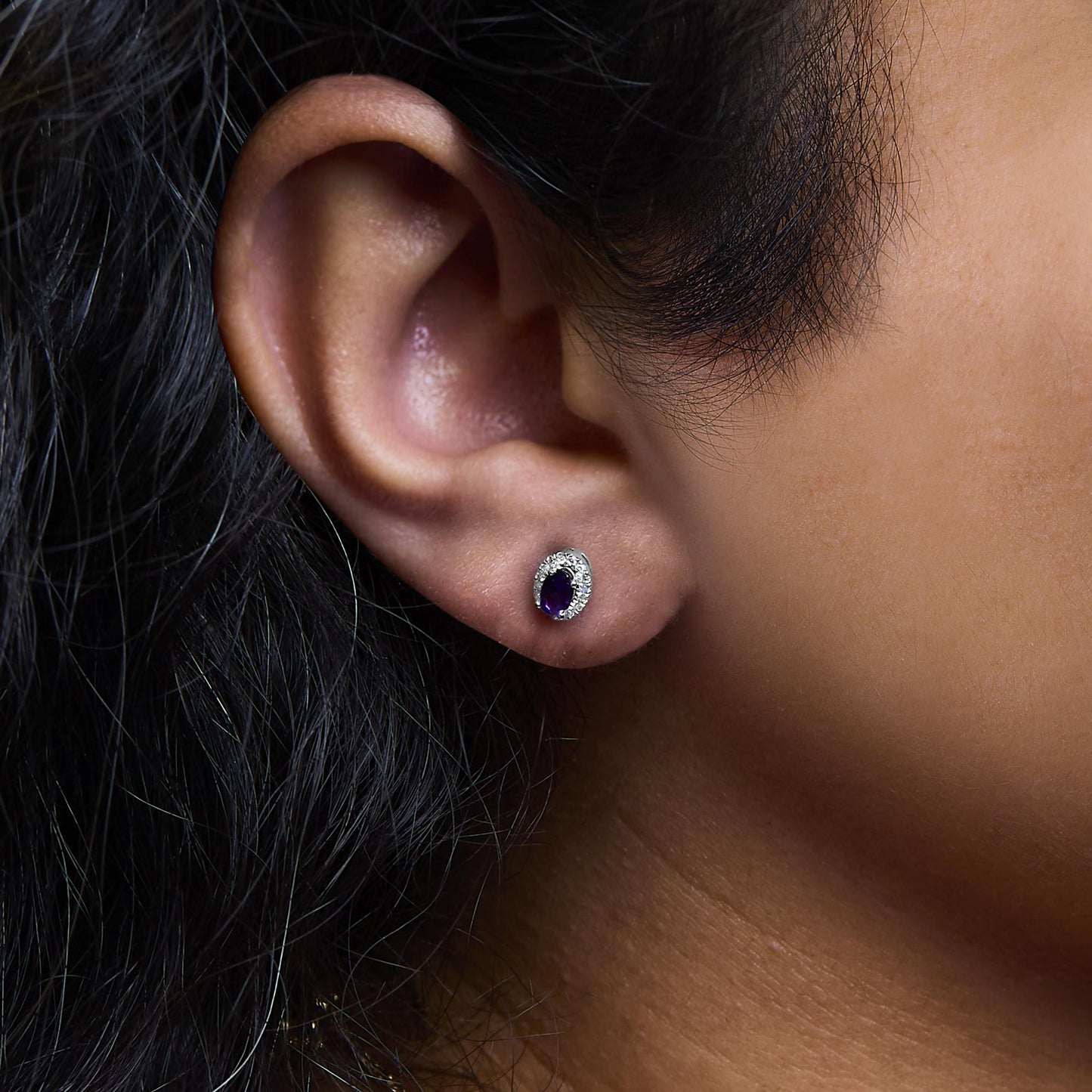 Close-up side view of a white metal earring with an oval purple gemstone surrounded by small white gemstones, worn on a model's ear.