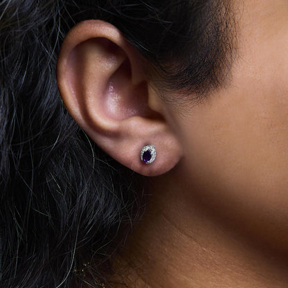 Close-up side view of a white metal earring with an oval purple gemstone surrounded by small white gemstones, worn on a model's ear.