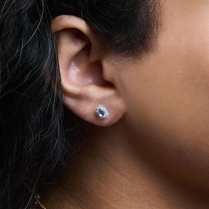Close-up side view of white metal stud earrings on a model’s ear, featuring oval blue gemstones surrounded by a cluster of small white gemstones in a pave setting.