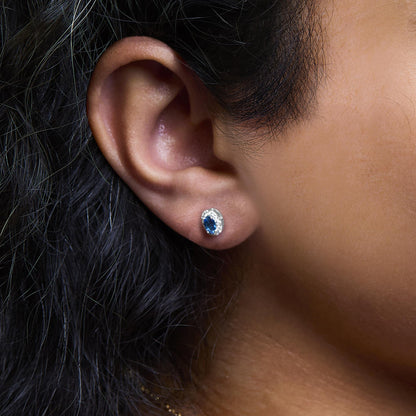 Close-up side view of a white metal stud earring on a model’s ear, featuring an oval blue gemstone surrounded by small white gemstones in a pave setting.