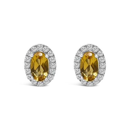 Pair of white metal stud earrings with oval yellow gemstones in four-prong settings, surrounded by a halo of small round white gemstones, shown from a front angle.