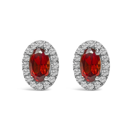 Front view of white metal oval earrings featuring a red oval gemstone center surrounded by a halo of small round white gemstones in pave setting.