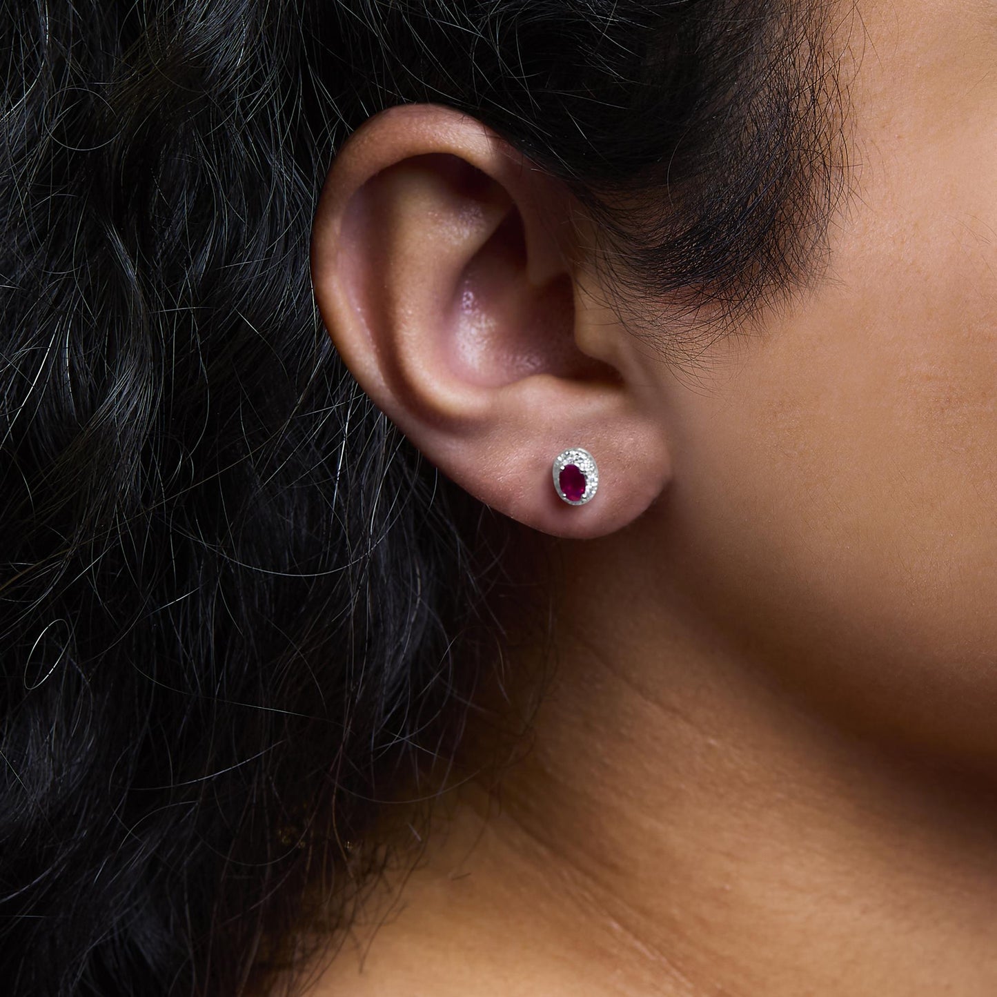 Close-up side view of a white stud earring on a model's ear featuring an oval red gemstone surrounded by small round white gemstones in a pave setting.