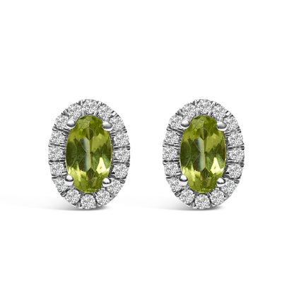 Front-facing shot of a pair of white metal oval earrings with green oval gemstones surrounded by a halo of small round white gemstones in pave setting.