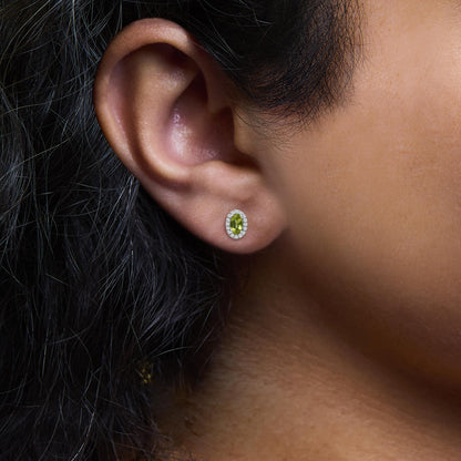 Close-up side view of a white metal earring on a model’s ear, featuring an oval green gemstone surrounded by small white gemstones in a pave setting.
