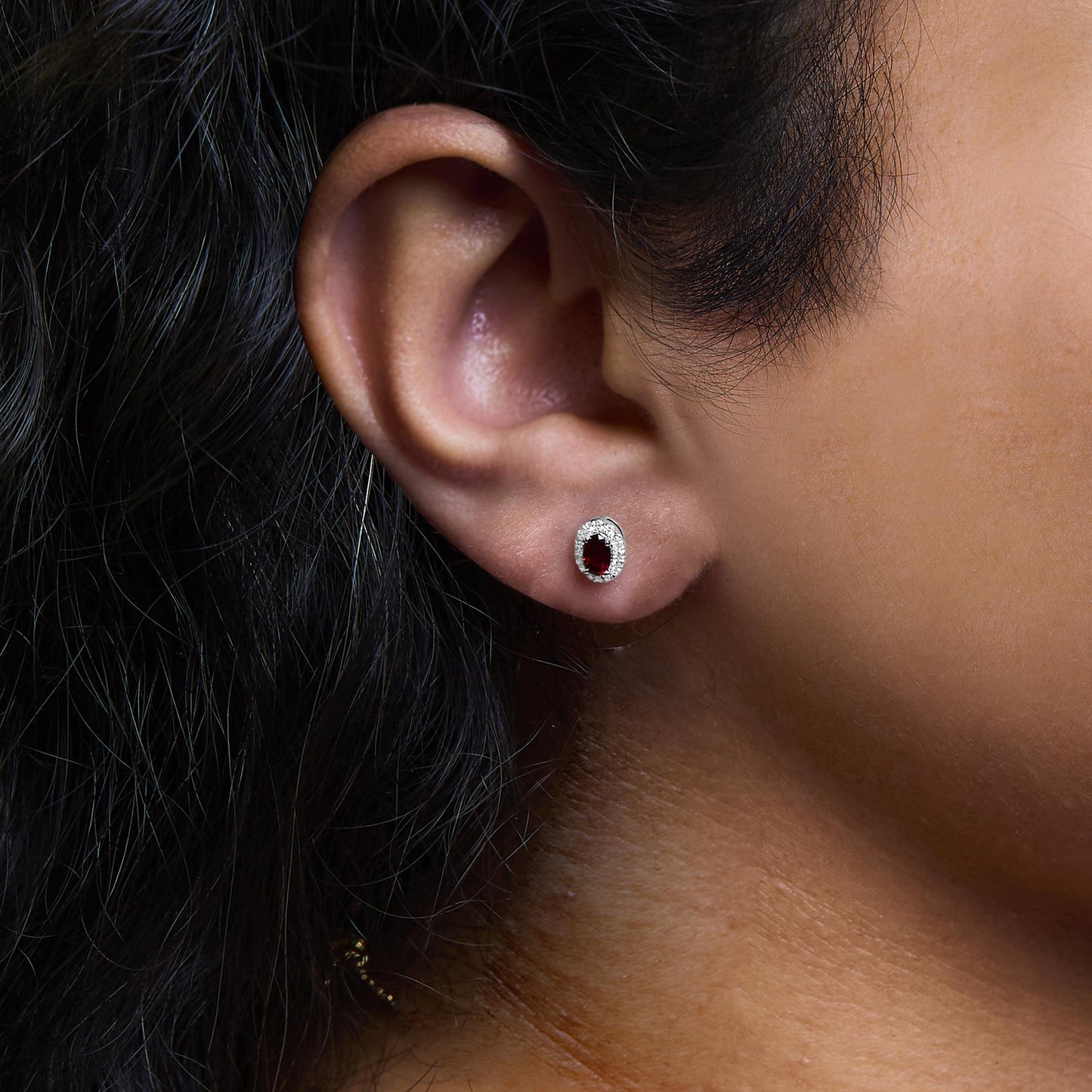 Close-up side view of a white metal earring worn on a model's ear featuring an oval red gemstone prong-set in the center, surrounded by a halo of small round white gemstones in a pave setting.