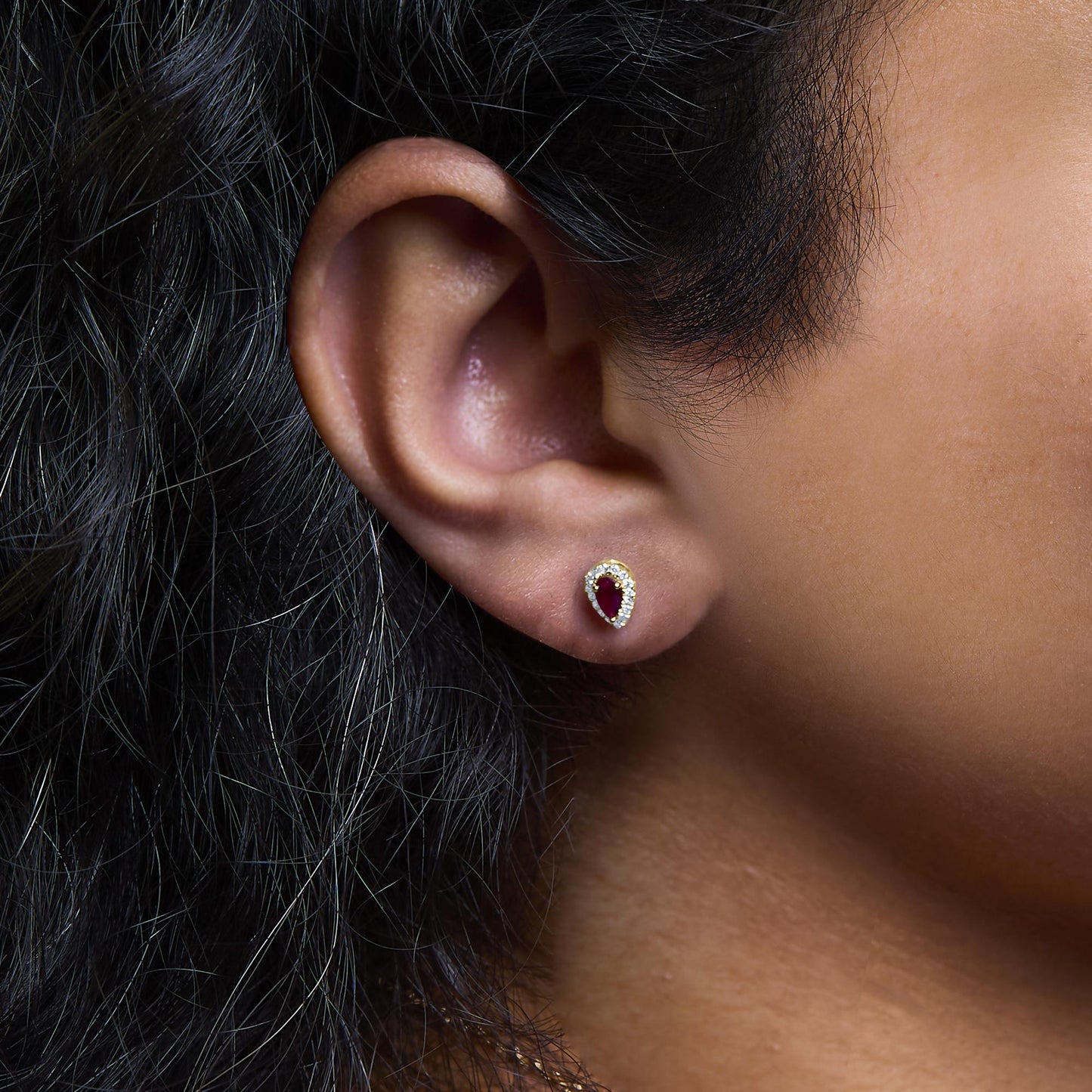 Close-up side view of a yellow metal earring on a model's ear featuring a pear-shaped red gemstone surrounded by small white gemstones in a pave setting.