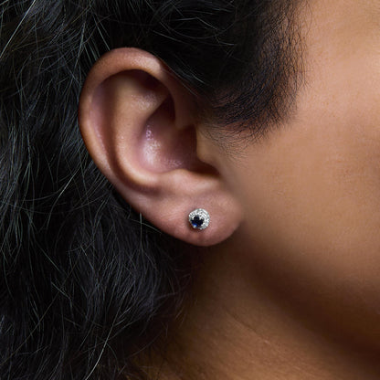 Close-up side view of a white metal stud earring on a model’s ear with a central blue oval gemstone surrounded by small round white gemstones in a pave setting.