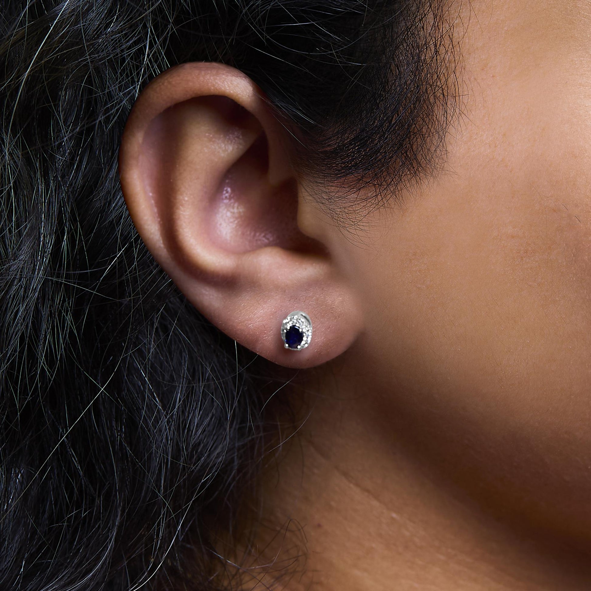 Close-up profile view of a white metal earring on a model's ear featuring a blue gemstone surrounded by small white gemstones in a pave setting.