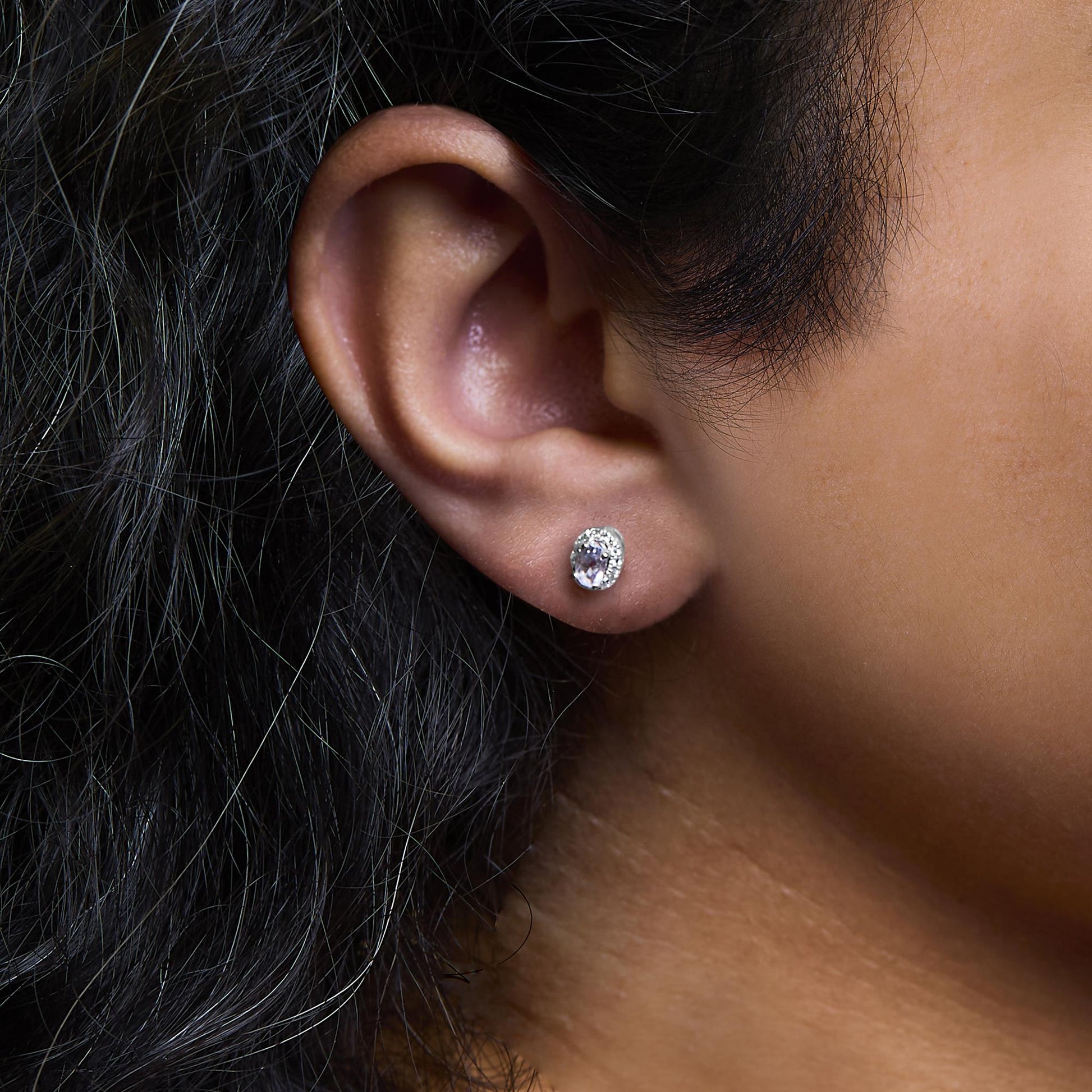 Close-up side view of an earlobe on a model wearing a white metal stud earring with an oval white gemstone surrounded by small round white gemstones in a pave setting.
